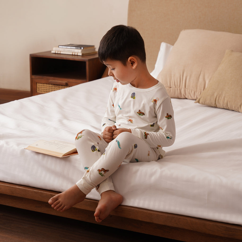 Child in long pajamas with animal print sitting on a bed reading a book in a cozy bedroom setting.