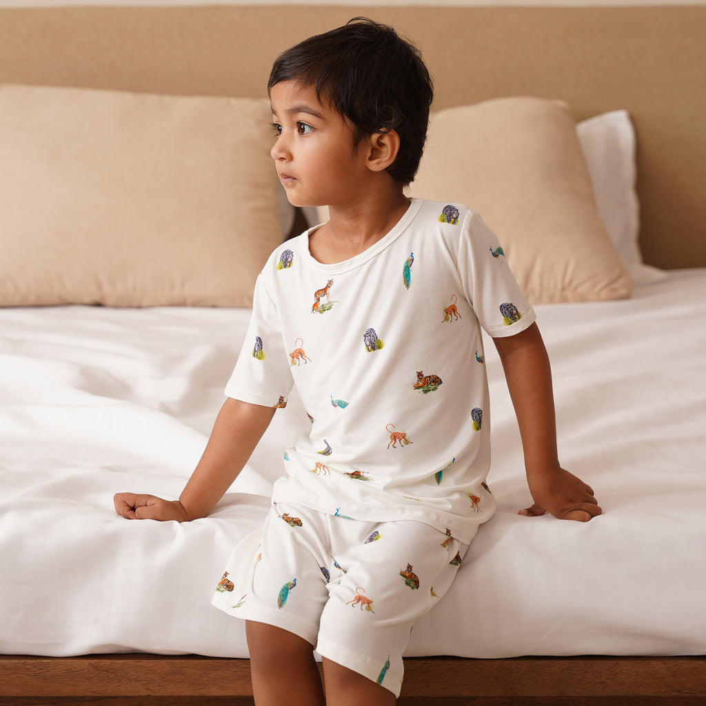 Child wearing a white short pajama set with animal prints sitting on a bed.
