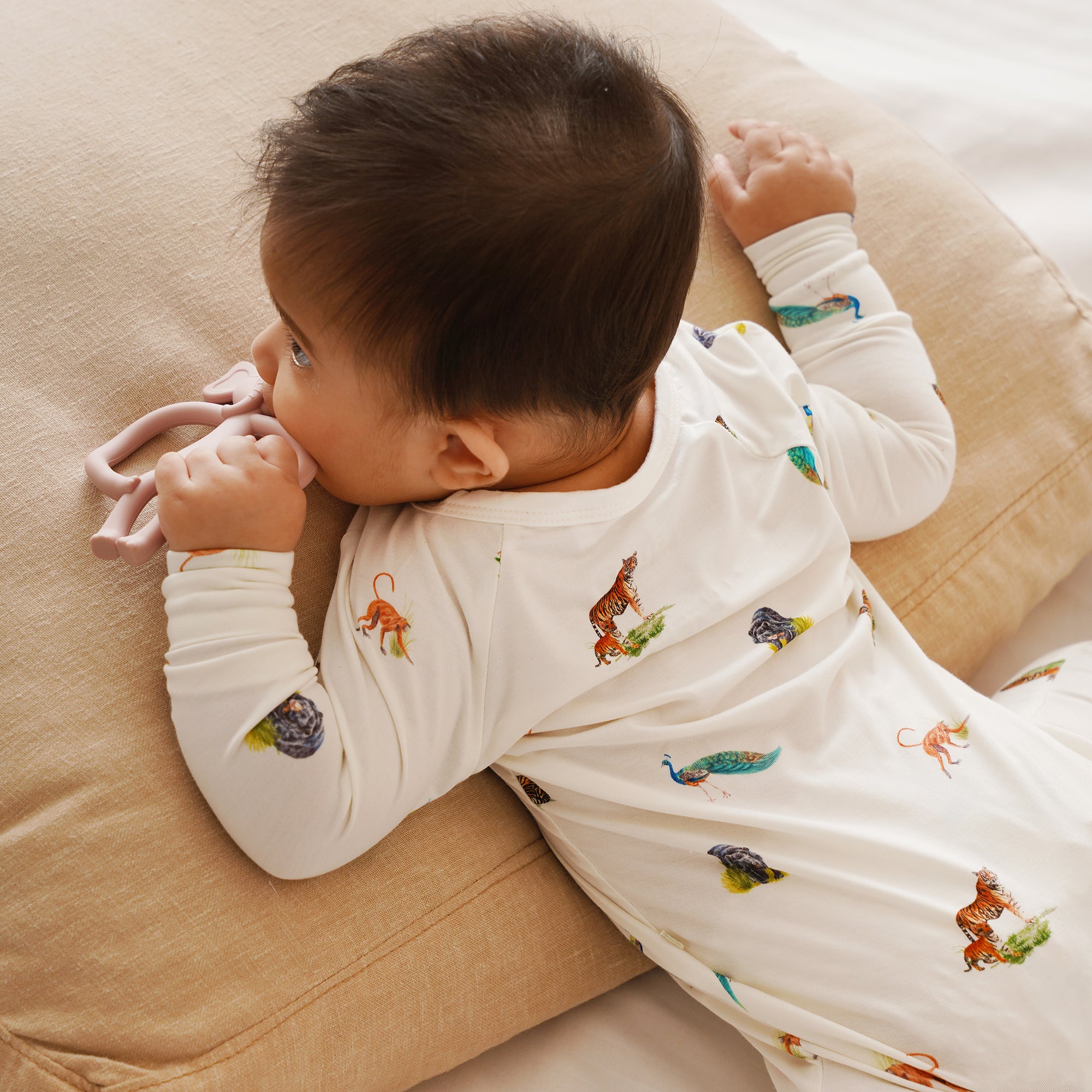 Baby wearing a white onesie or zipsuit with colorful animal prints, lying on a beige surface.