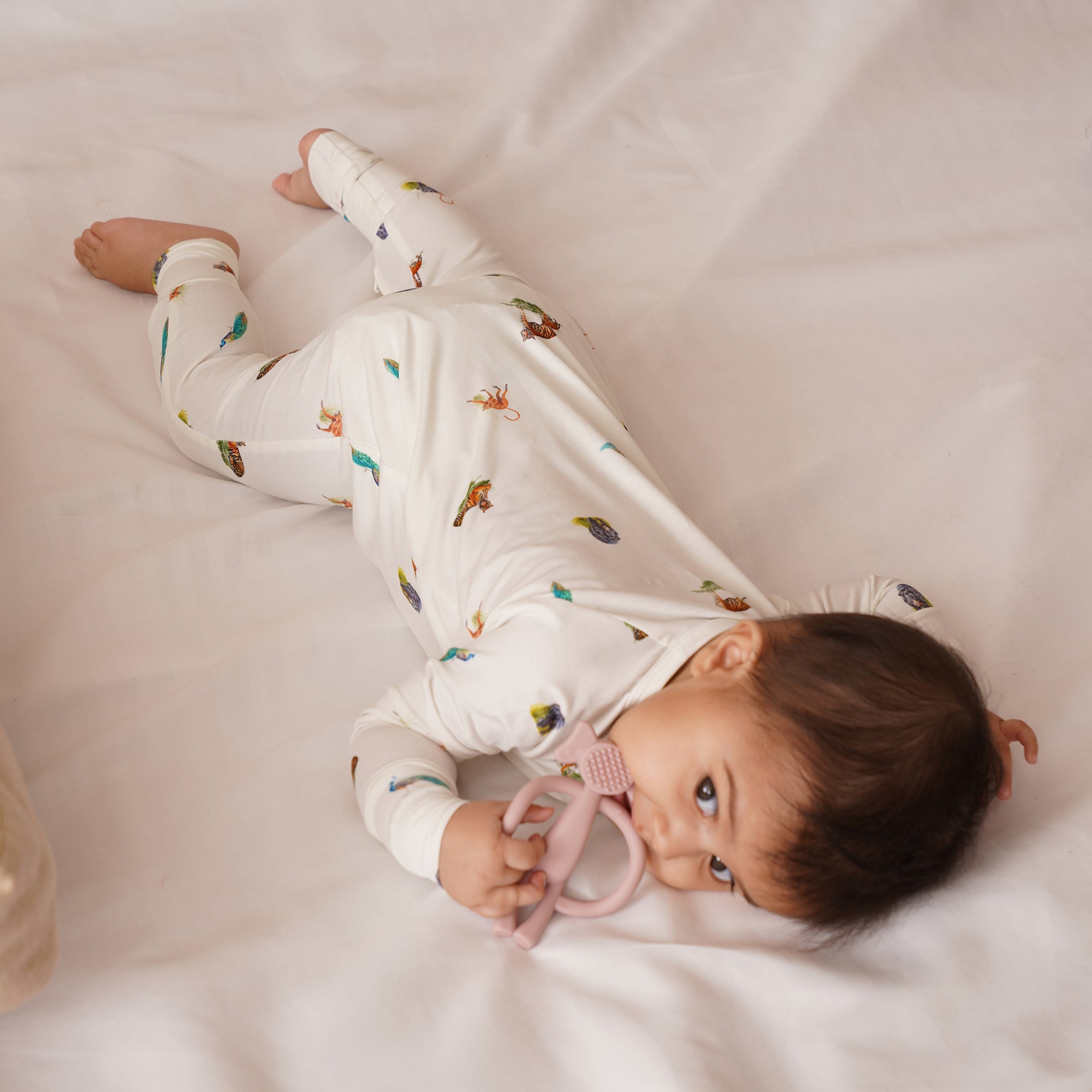 Baby lying in animal print zipsuit on a white blanket holding pink teether