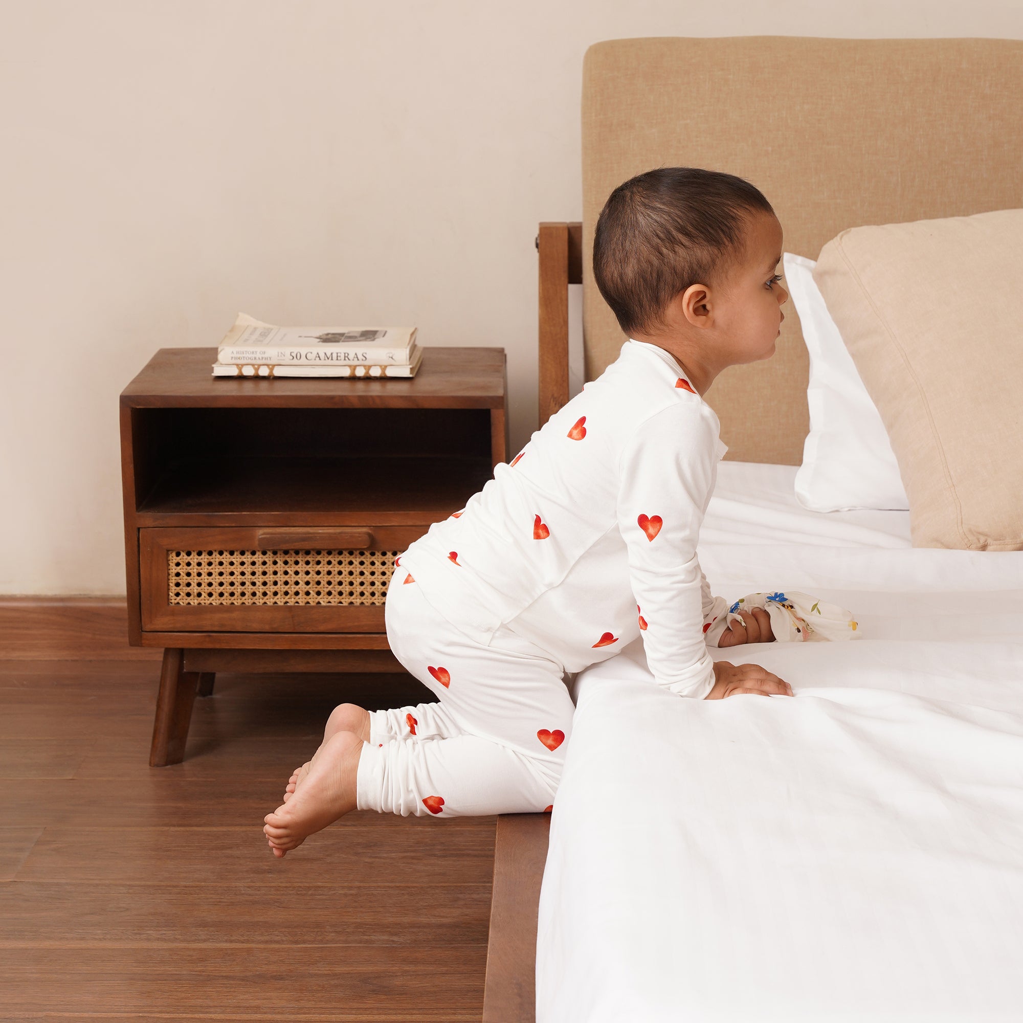 Child in a white onesie with red hearts sitting on the floor next to a bed.
