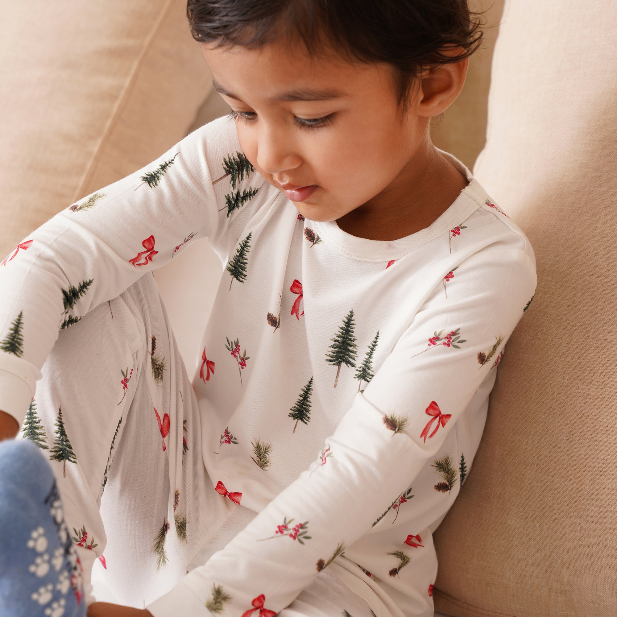 Child wearing a white long-sleeve shirt with Christmas tree and bow pattern, sitting on a beige couch.