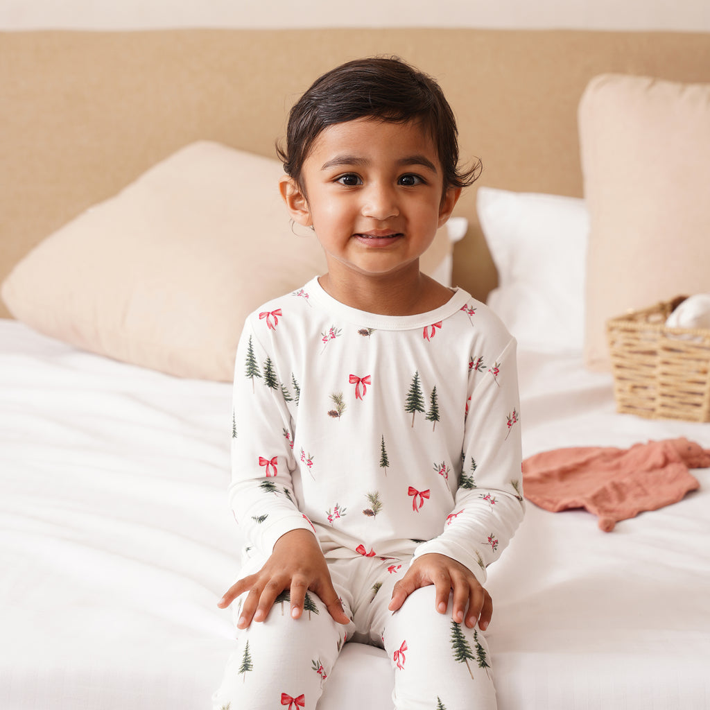 Child wearing pajamas with a Christmas tree and bow pattern, sitting on a bed.