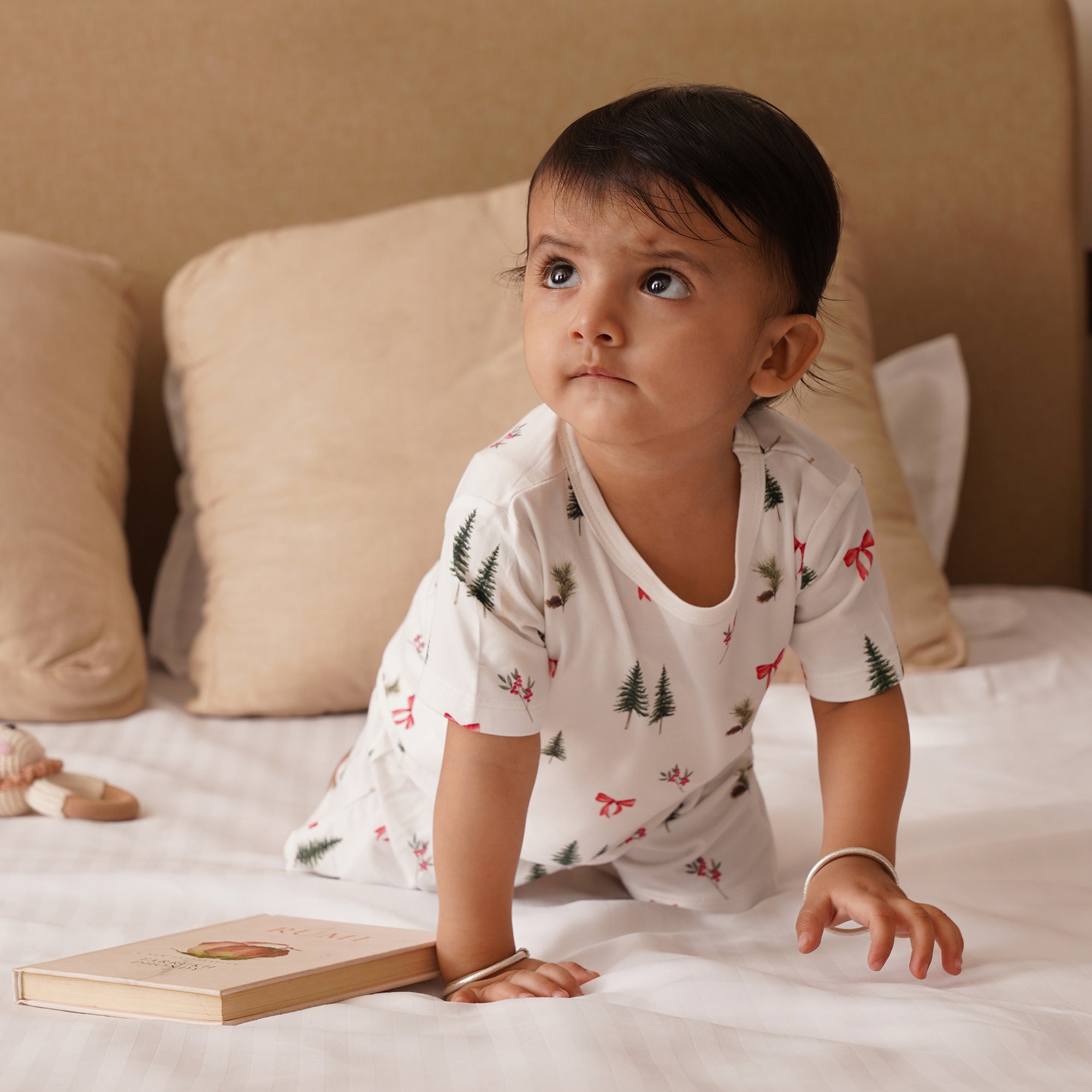 Baby sitting on a bed holding a wooden book in holiday print short pjs