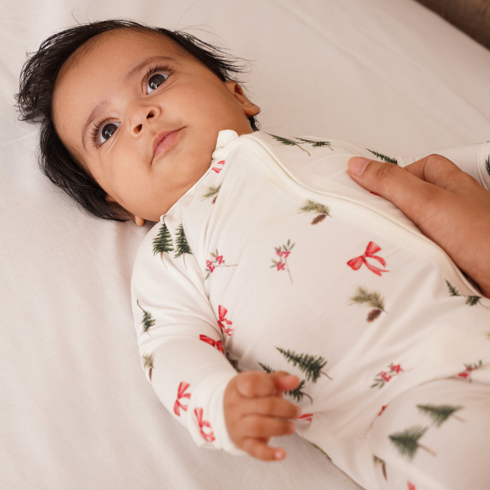 Baby wearing a white onesie or zipsuit with green tree patterns for holidays, lying on a white surface.