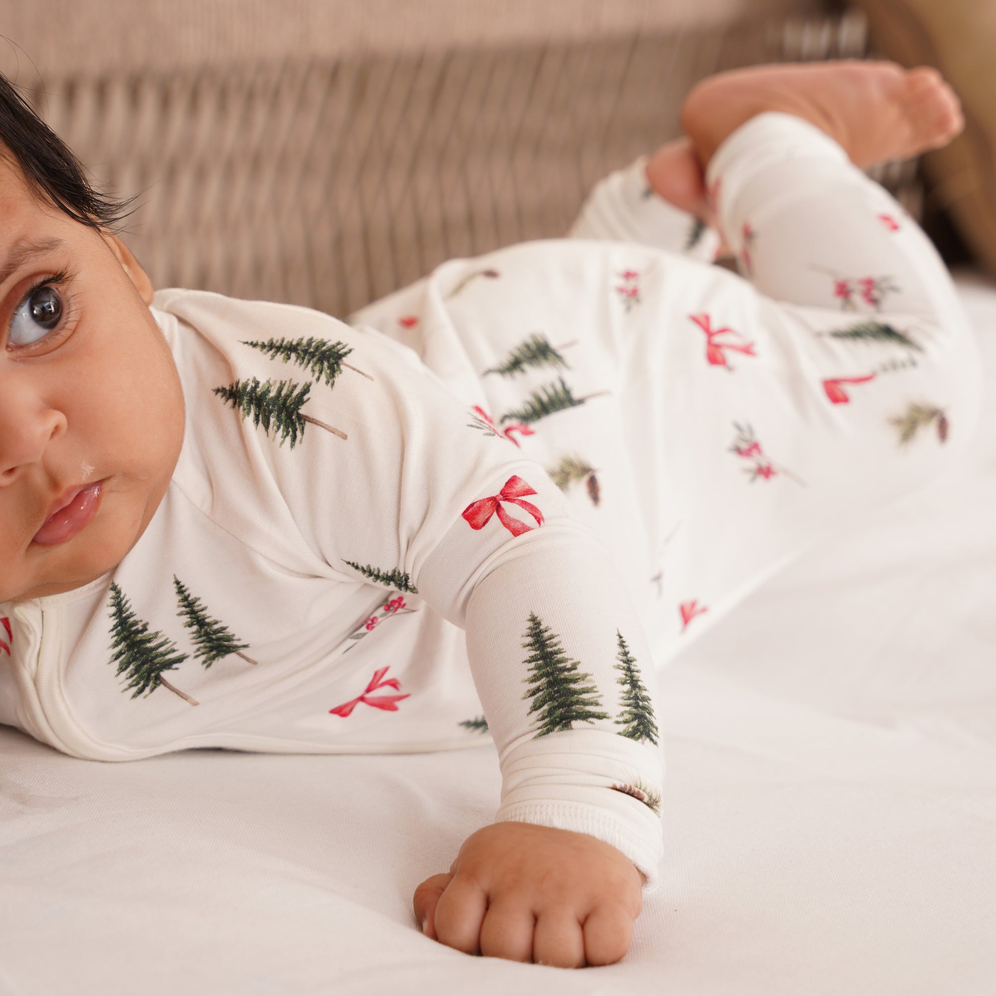 Baby wearing a white onesie with Christmas tree and bow pattern, lying on a bed.