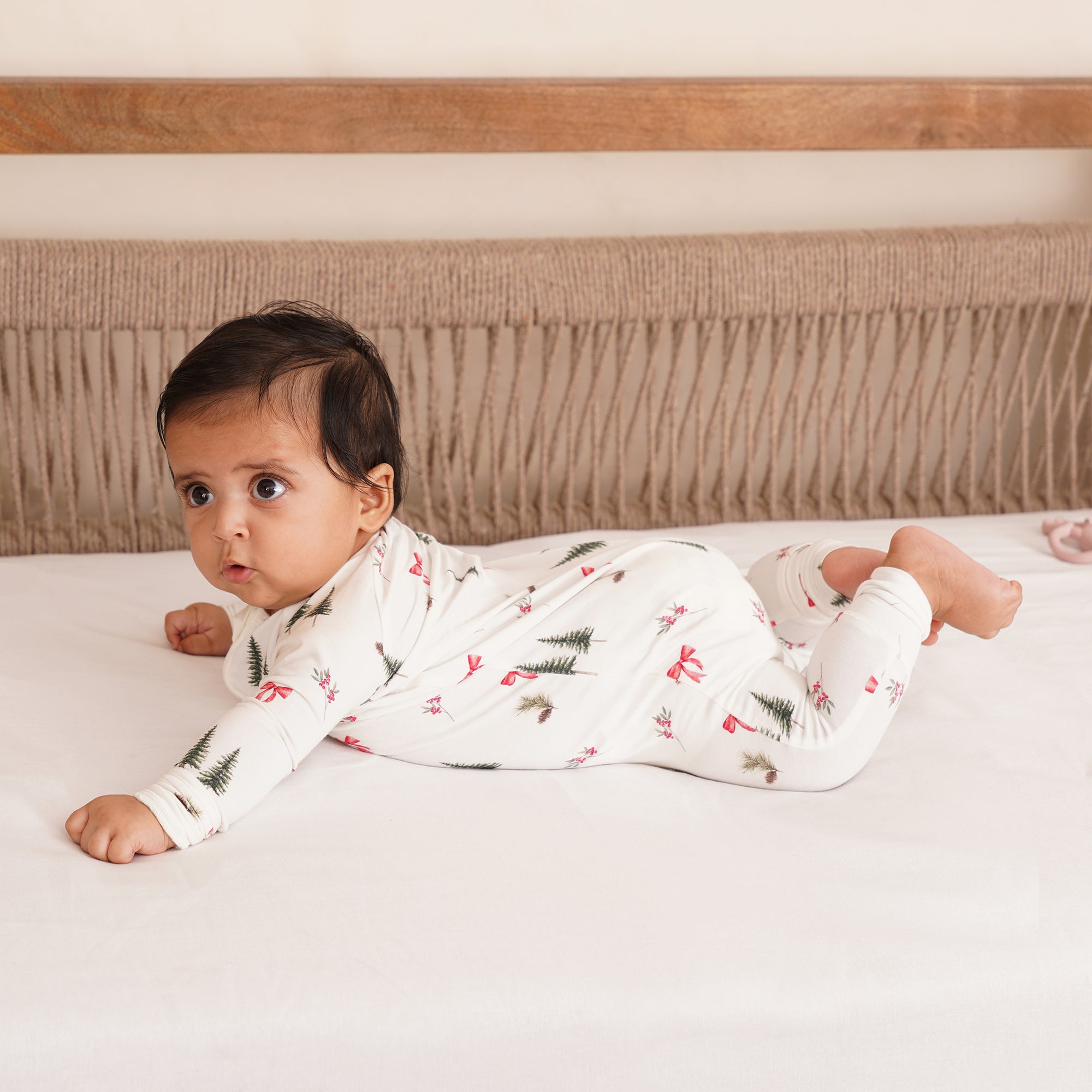 Baby lying on a bed wearing a white onesie or zipsuit with red and green patterns for holiday season.