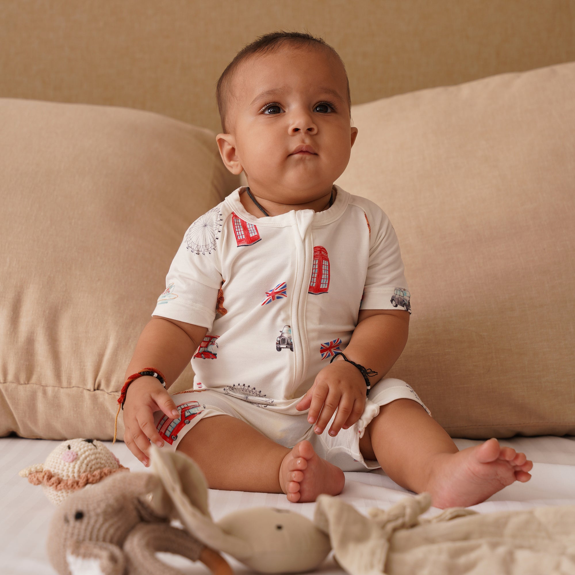 Baby sitting on a couch with toys around, wearing a white onesie with red and blue designs london themed