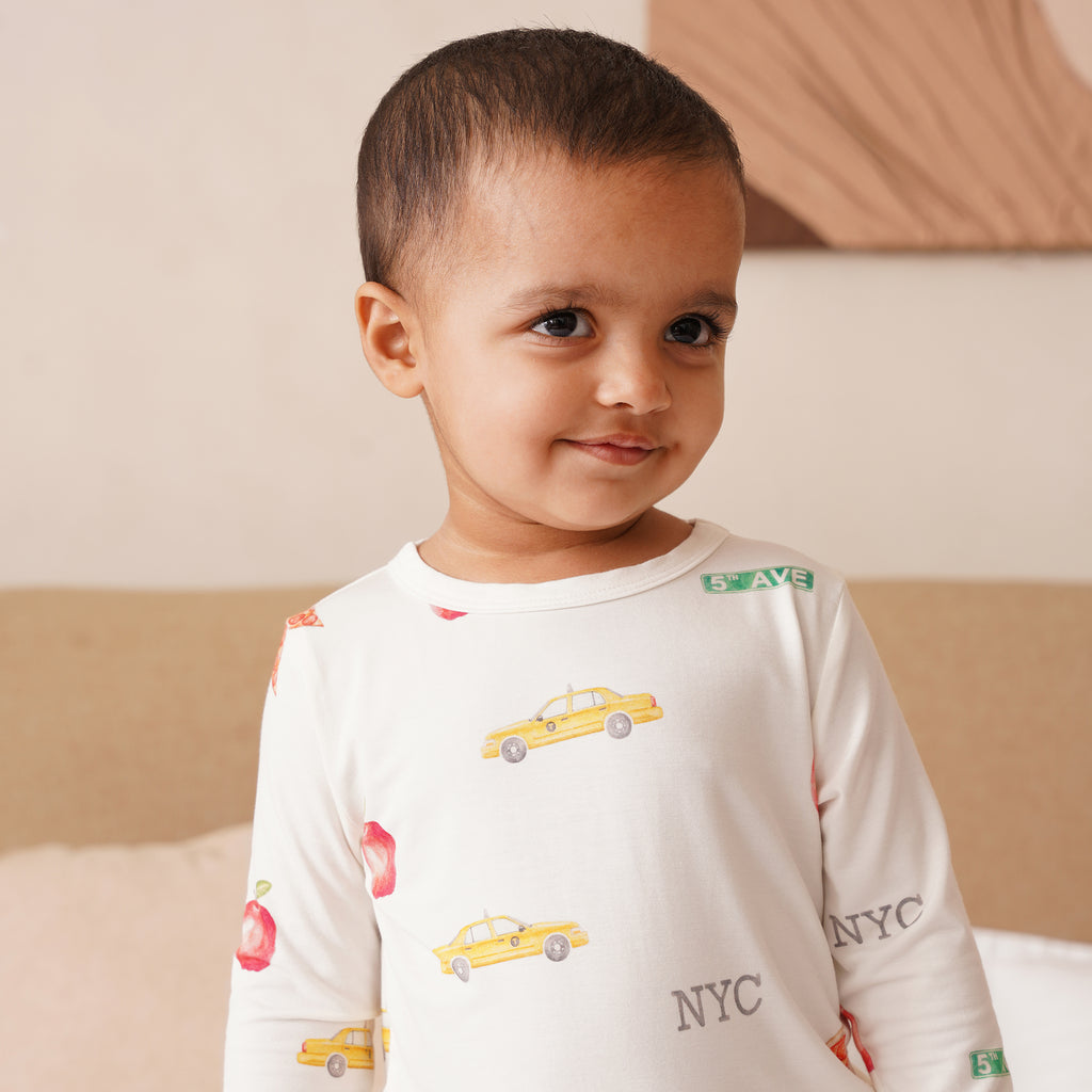 Child wearing a long-sleeve shirt with taxi and 'NYC' print, sitting on a bed.
