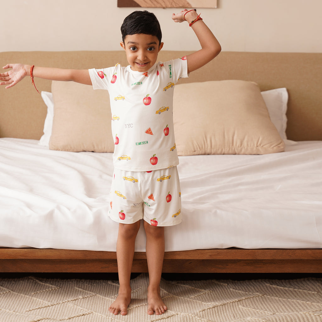 Child wearing a  short pajama set with NYC patterns standing in front of a bed.