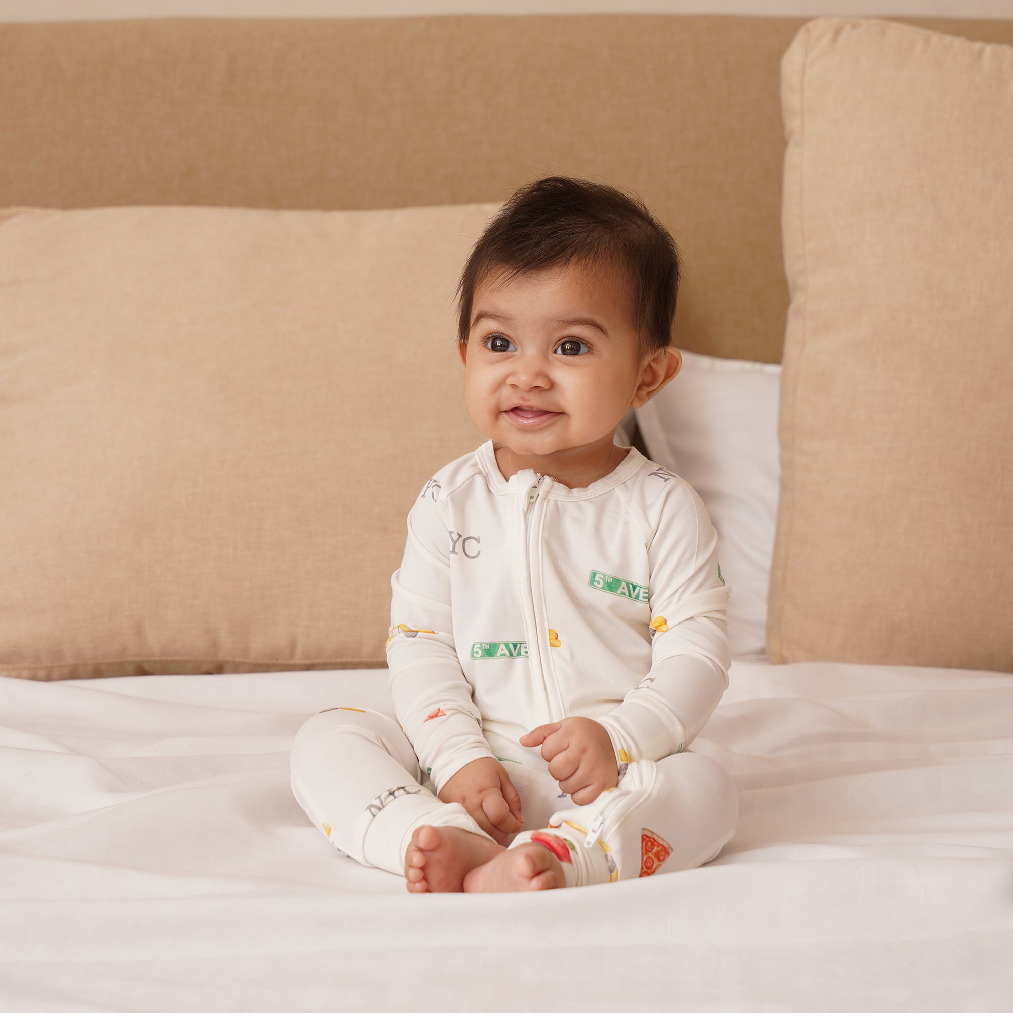 Baby sitting on a bed wearing a white onesie with colorful NYC themed patterns.