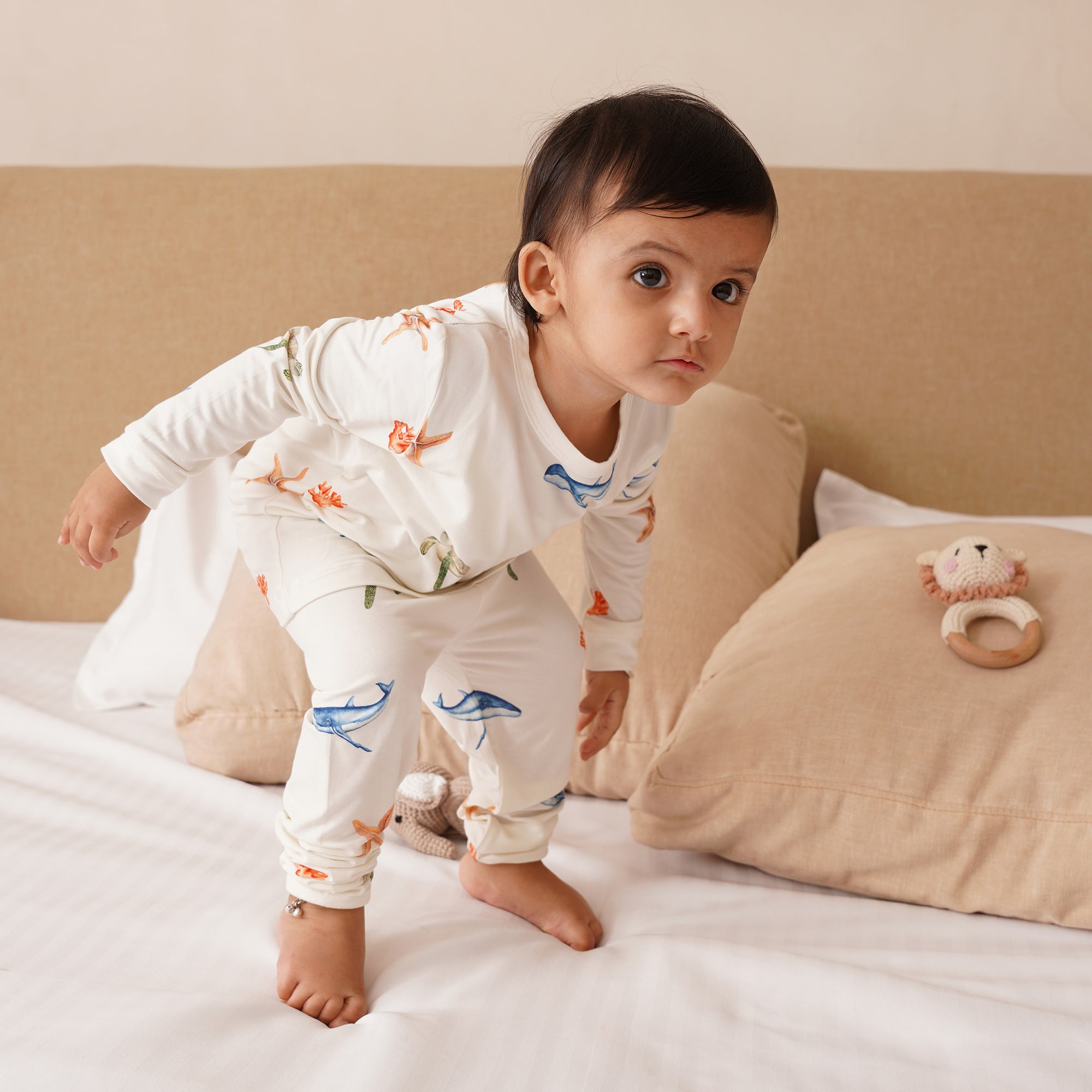 Baby in long pajamas under the sea patterned standing on a bed with a toy nearby