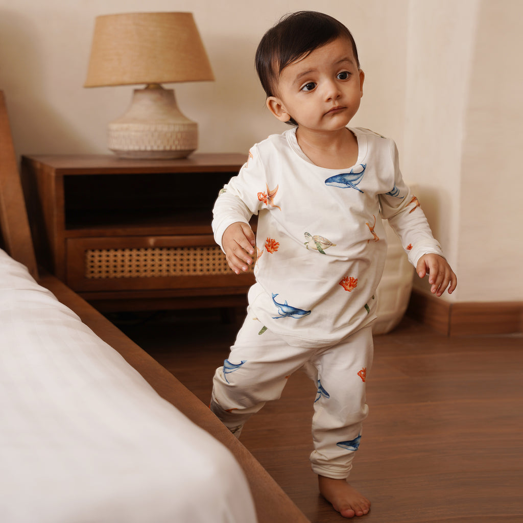 Baby in a white outfit long pajamas with colorful under the sea designs standing in a bedroom.