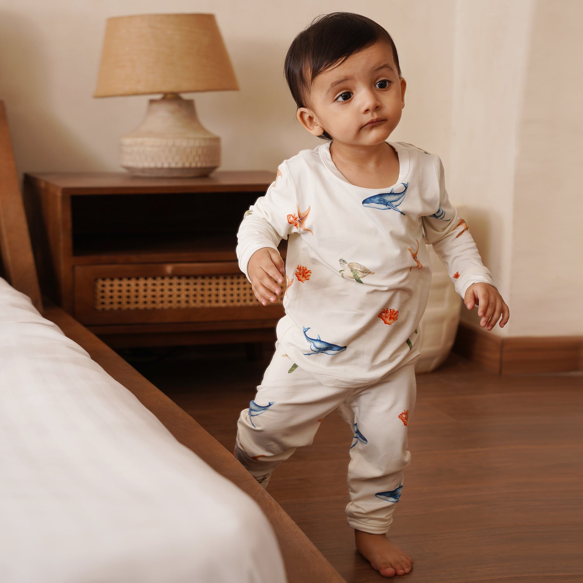 Baby in a white outfit long pajamas with colorful under the sea designs standing in a bedroom.