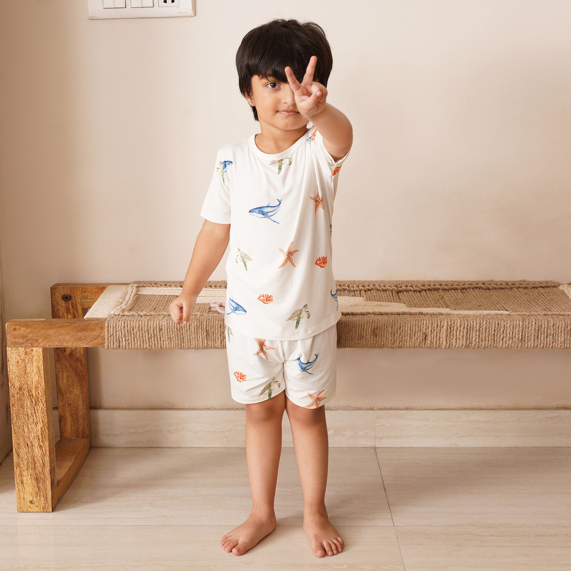 Child wearing a white outfit with colorful under the sea prints, standing in a room with a wooden bench.