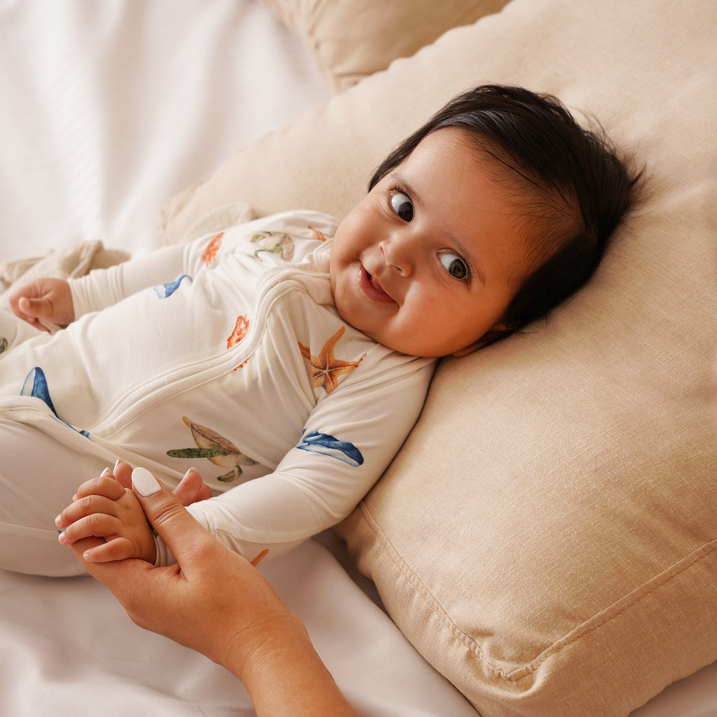 Baby lying on a beige pillow with a person holding their hand, wearing a white onesie with colorful under the sea patterns.